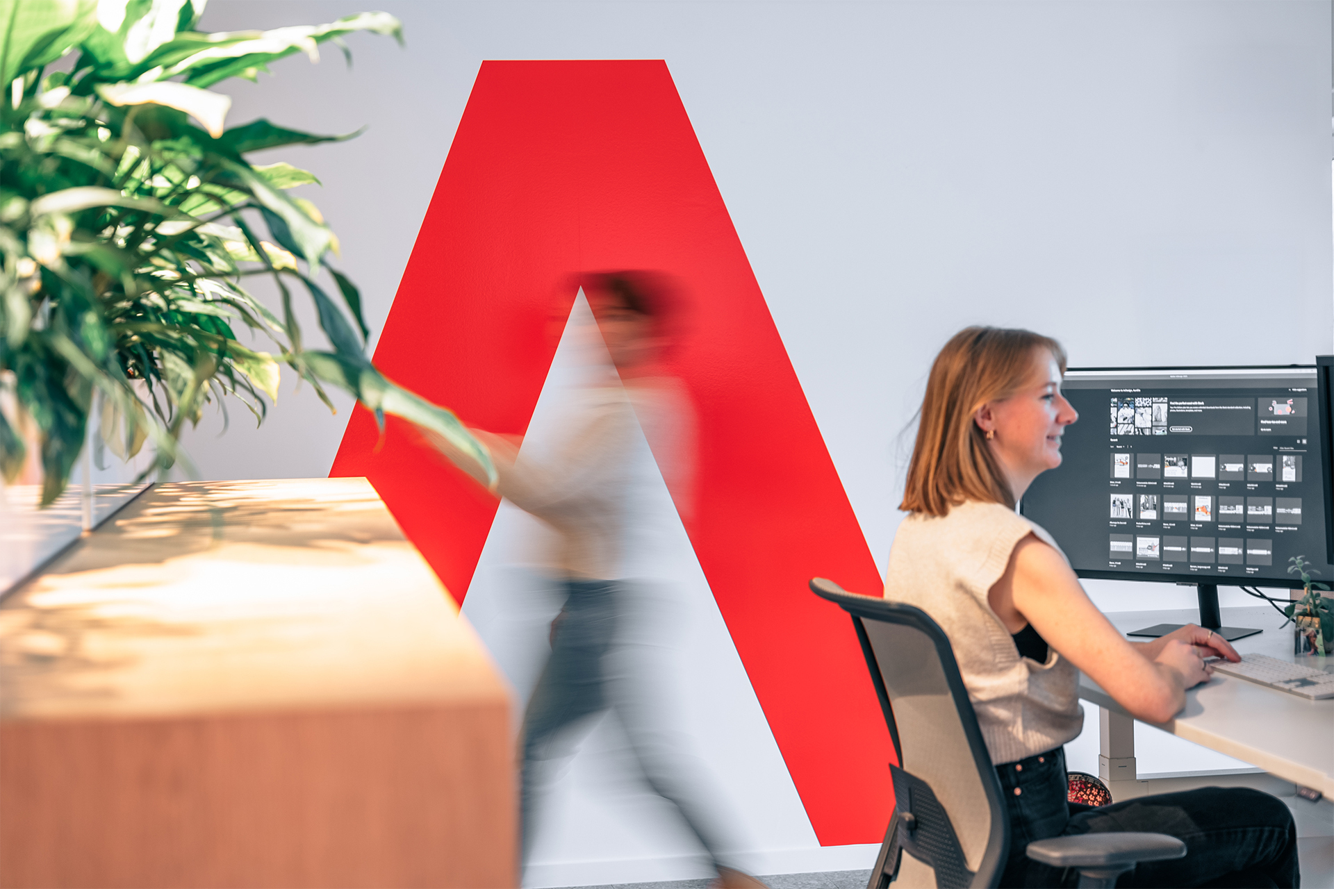 Medewerker aan het werk in een modern kantoor met groot rood A-logo op de muur en een collega die voorbij wandelt.