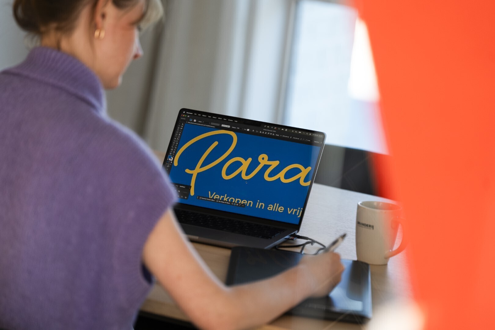 Vrouw werkt op een laptop met grafisch ontwerp op het scherm, waarop de tekst “Verkopen in alle vrijheid” te zien is. Naast haar staat een mok en een tekentablet.