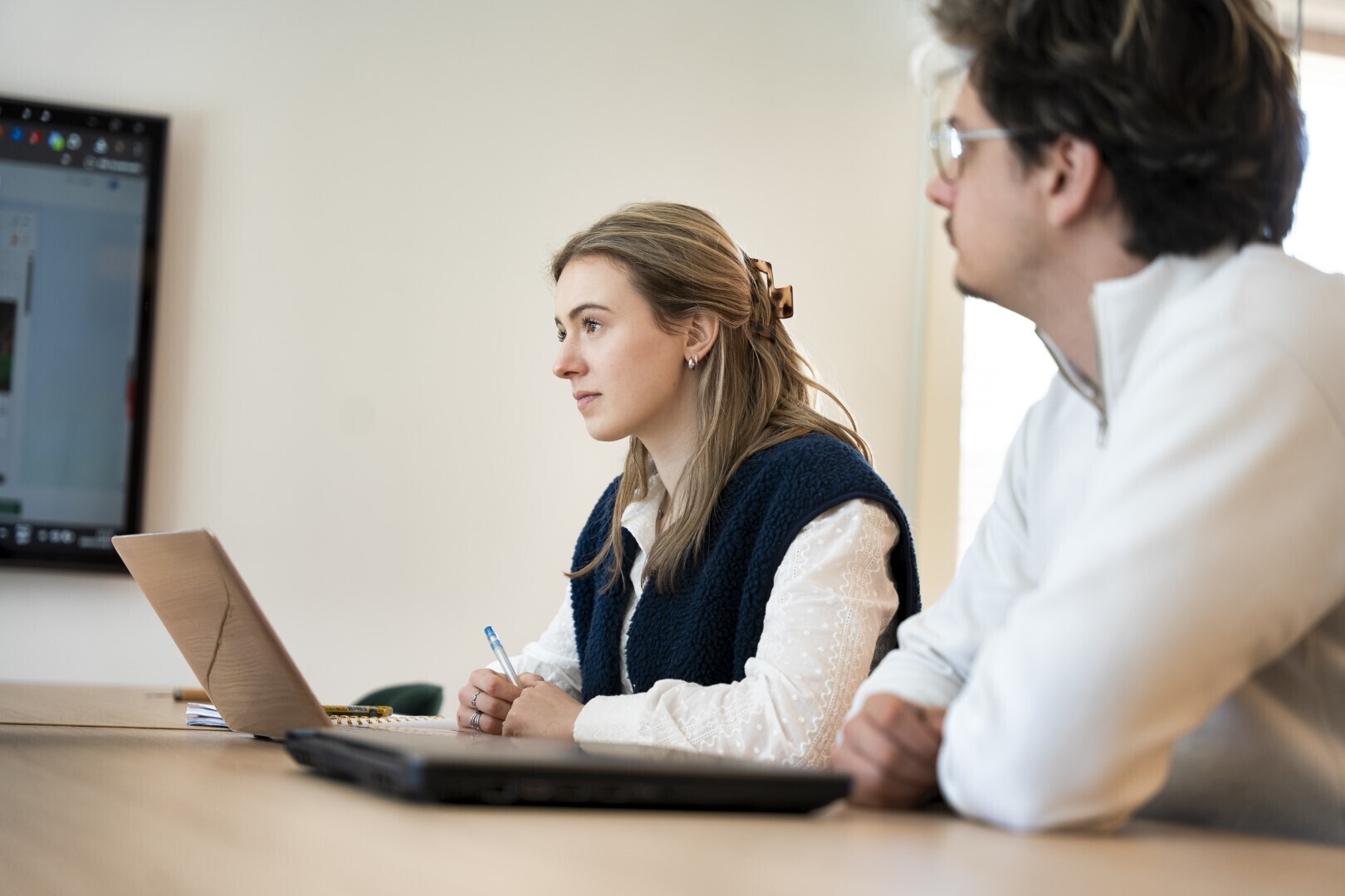 Twee collega’s aan een vergadertafel kijken aandachtig naar een persoon buiten beeld, met open laptops voor zich in een lichte vergaderruimte