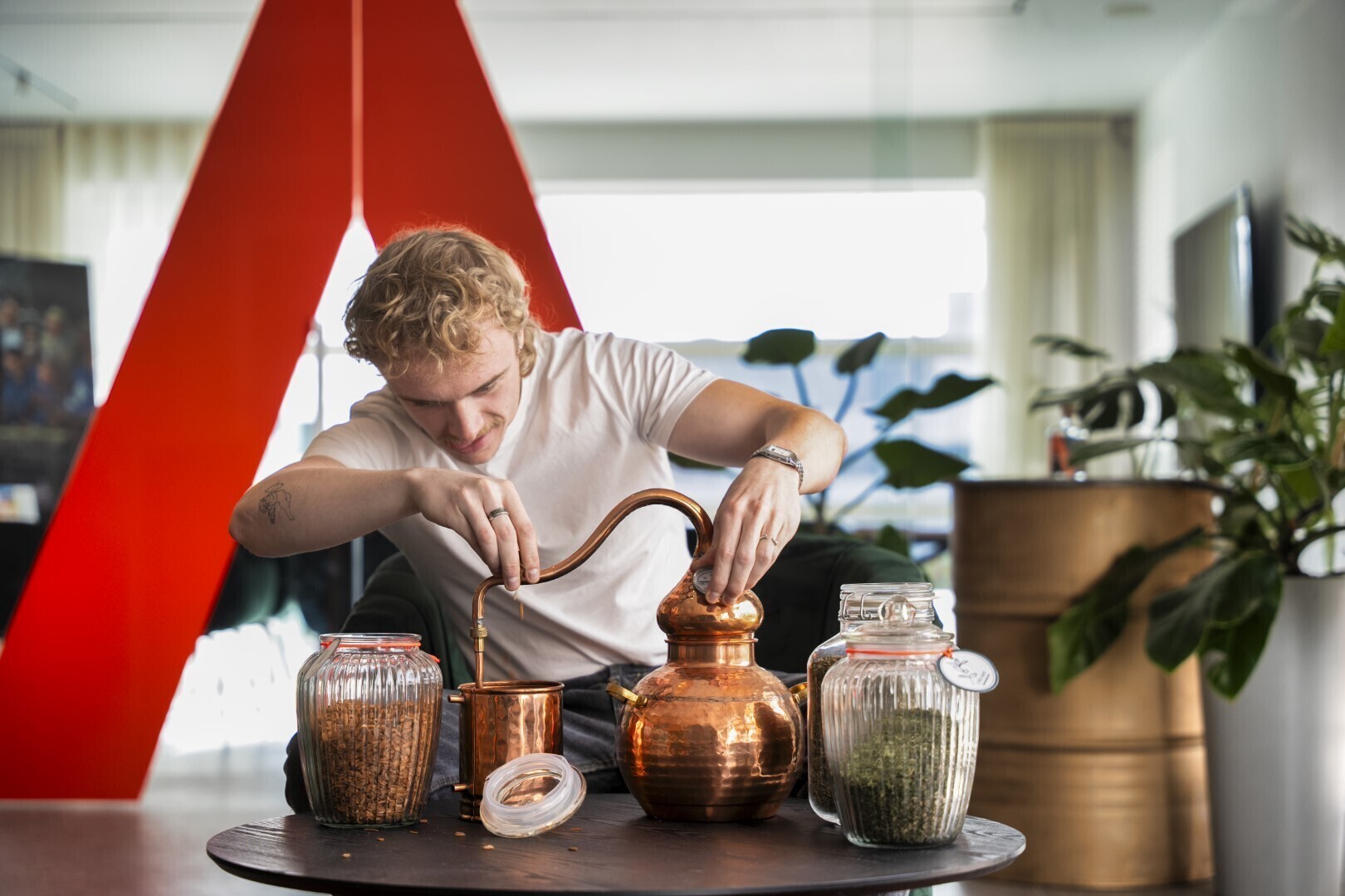 Man werkt met een koperen distilleerapparaat en potten met kruiden op een tafel in een modern kantoor met rode wand.