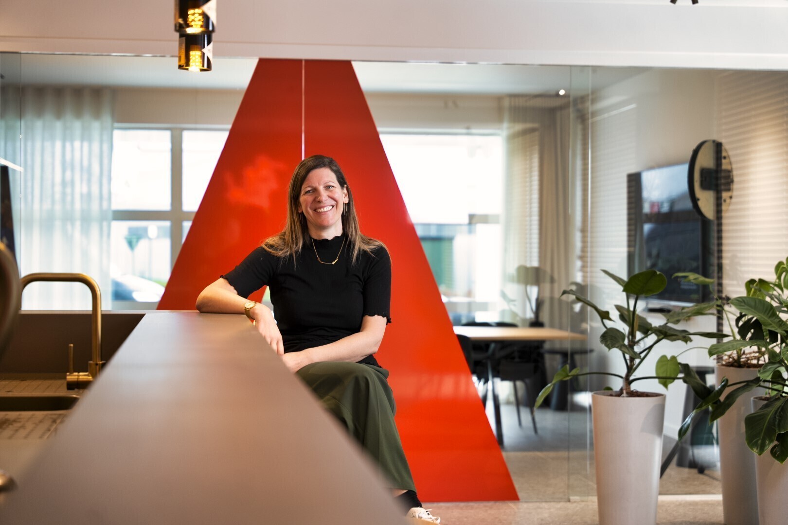 Vrouw zit lachend op een bureau in een modern kantoorinterieur met een opvallende rode wand op de achtergrond.