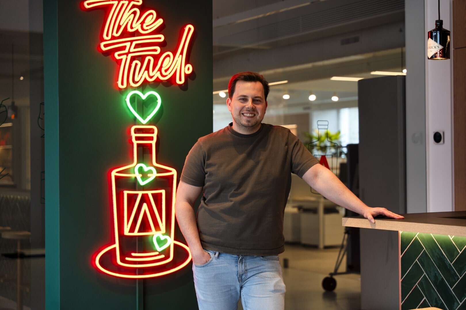 Man leunt ontspannen tegen een bar met op de achtergrond een neonillustratie van een fles en de tekst “The Fuel”.