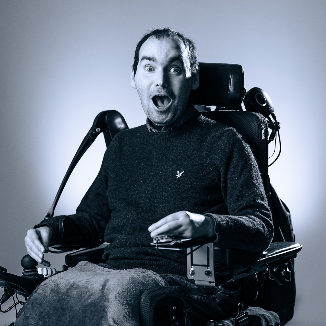 Lachende man in een elektrische rolstoel, gekleed in een donkere trui, poseert voor de camera tegen een neutrale achtergrond